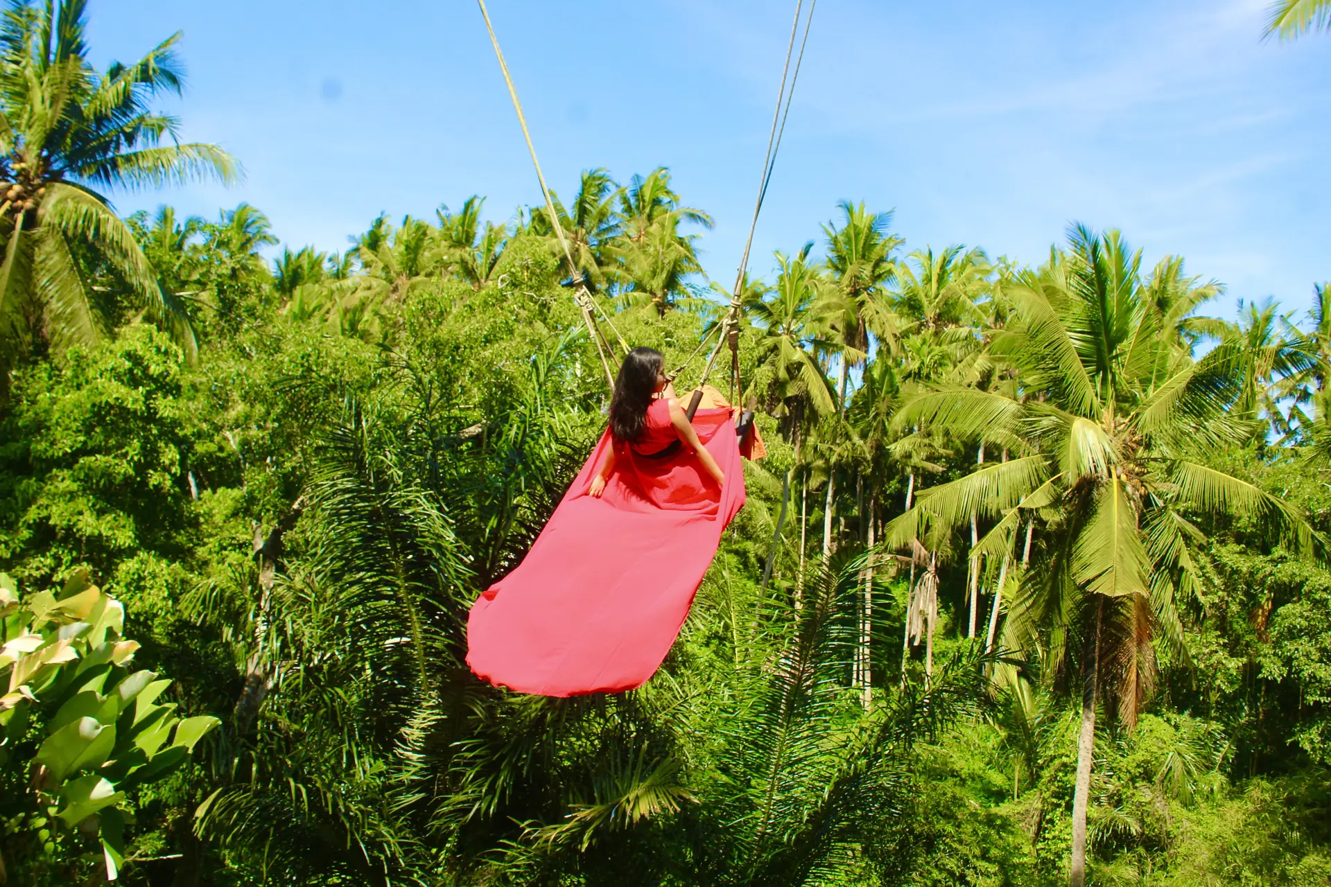 A woman in a long red dress on a rope swing high above palm jungle in Bali
