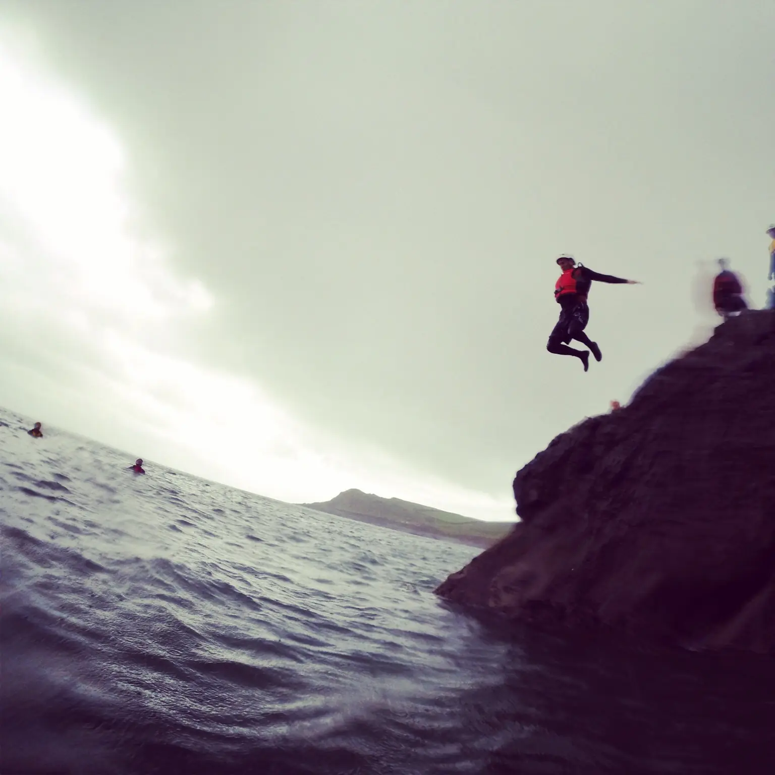 A coasteering jumper in a wetsuit and helmet mid-air off a dark rock ledge into the sea, others swimming below (symbolic image, Gower, not Mallorca)