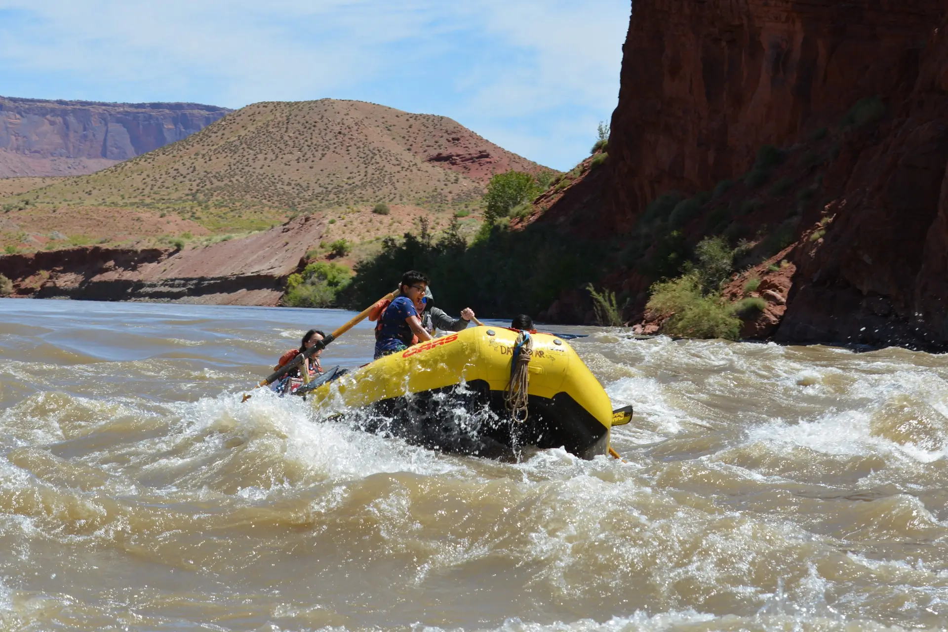A yellow raft dropping into a wave on the Colorado River below red cliffs