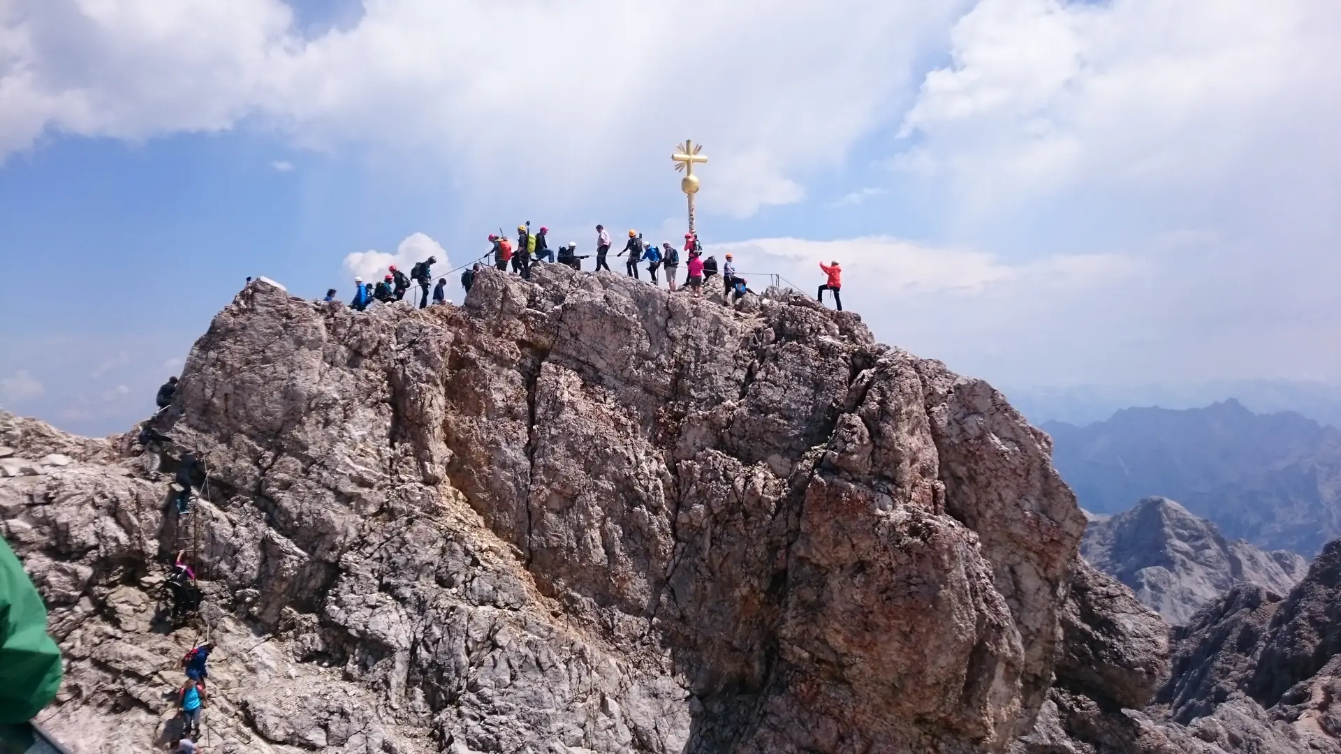 Das goldene Gipfelkreuz der Zugspitze mit Besuchern auf dem Felsgrat im Sommer