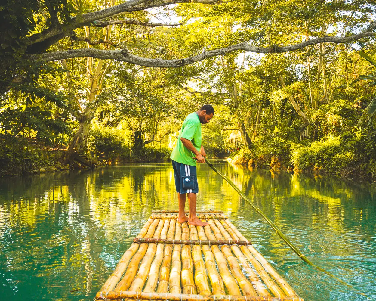 A raft captain poling a bamboo raft down the green, tree-lined Martha Brae River