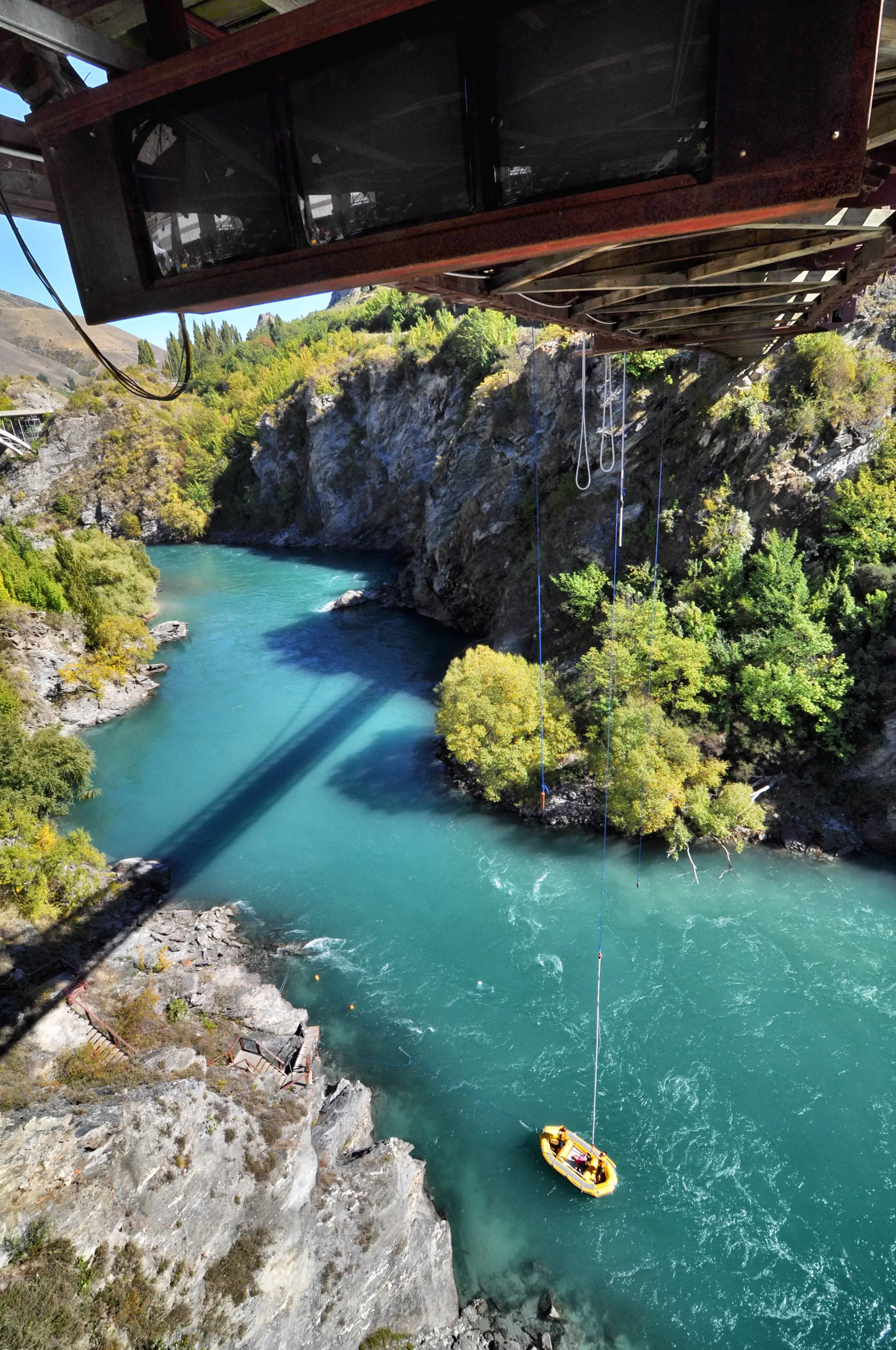 A bungy jumper hanging on the cord below the Kawarau Bridge above the turquoise river, a raft waiting on the water