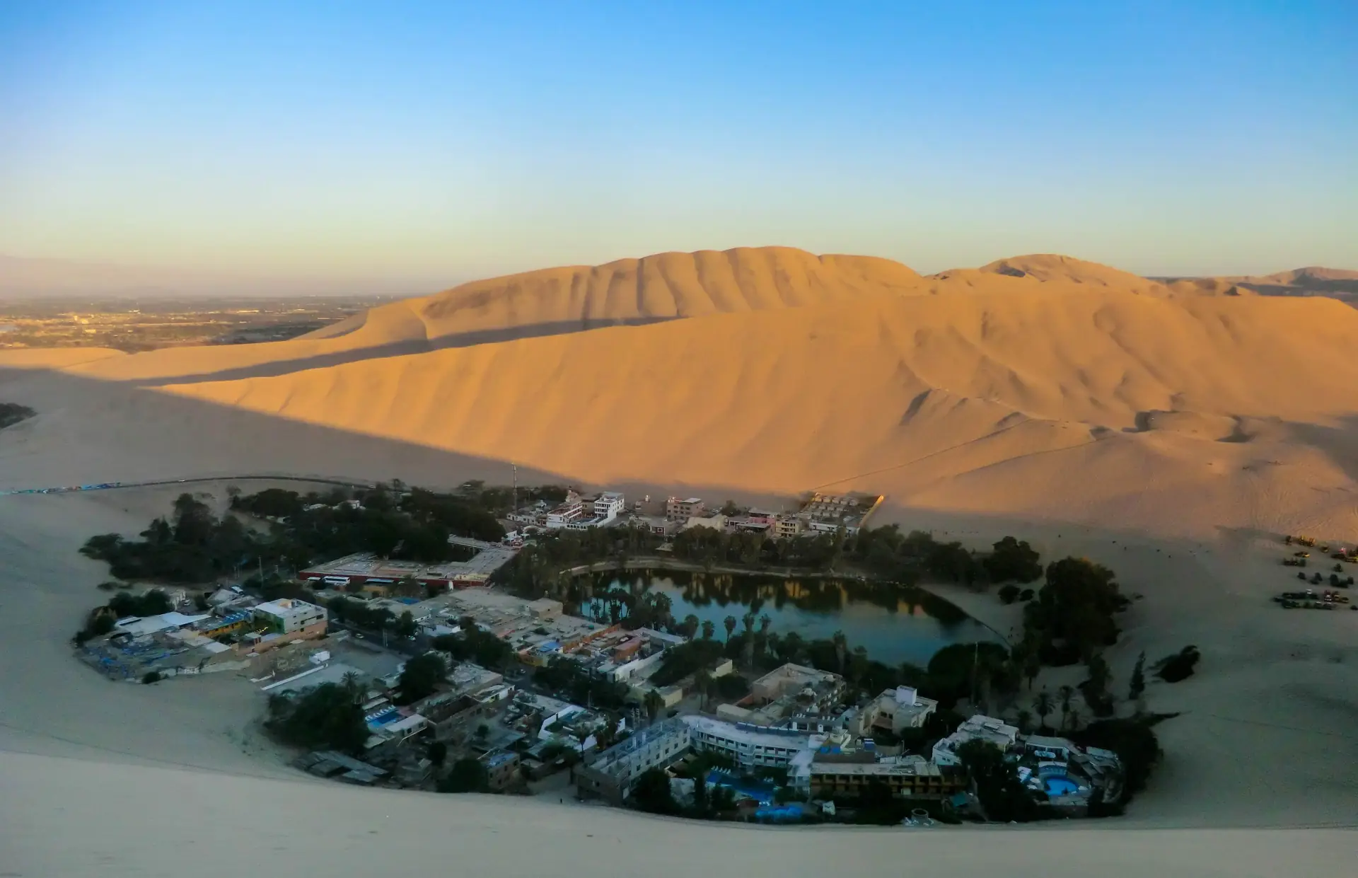 Die Oase Huacachina von oben: eine grüne Lagune, umringt von Palmen und Hotels, auf allen Seiten von hohen Sanddünen eingeschlossen