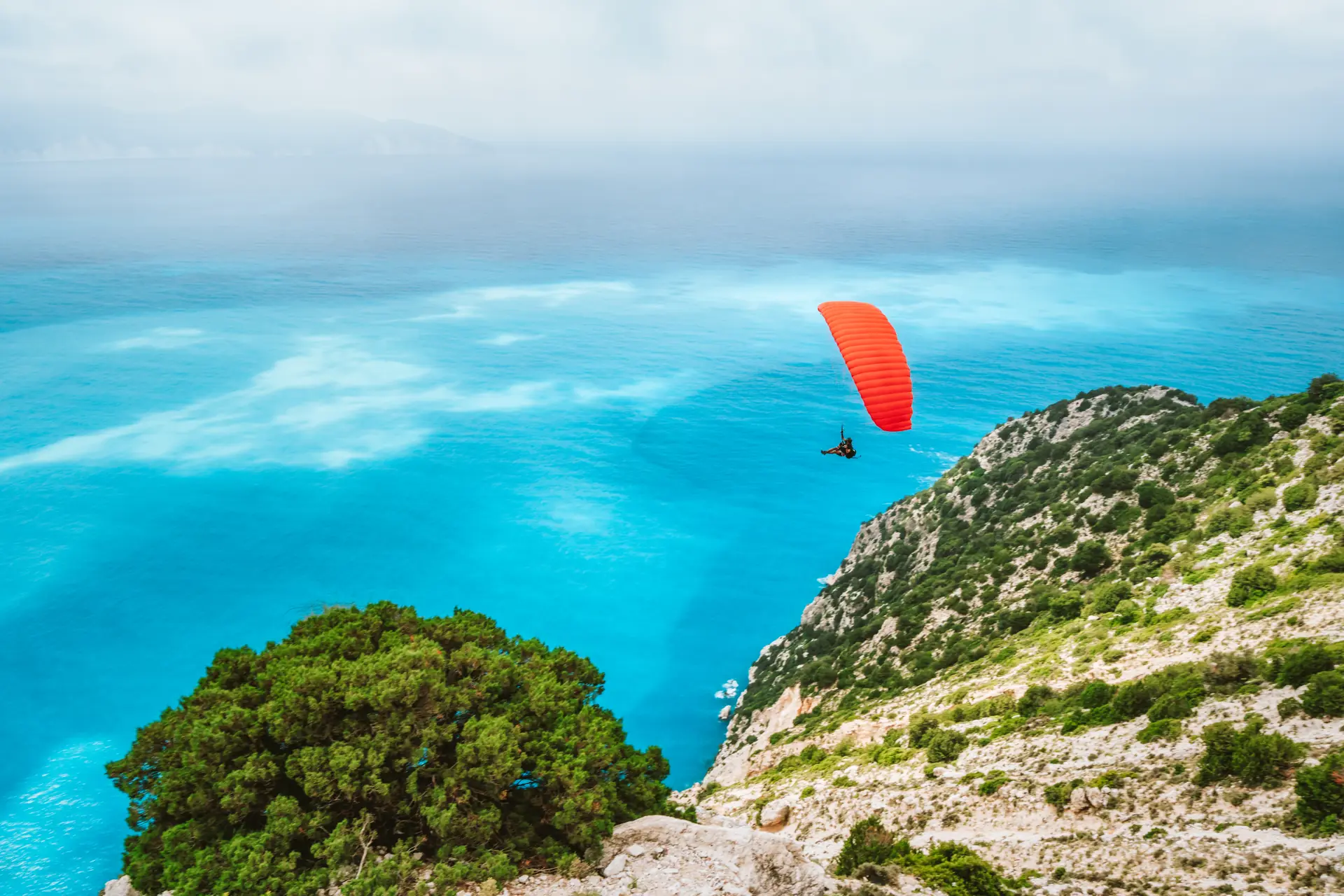 A red paraglider high above a turquoise bay and white cliffs (symbolic image, Kefalonia, not Budva)