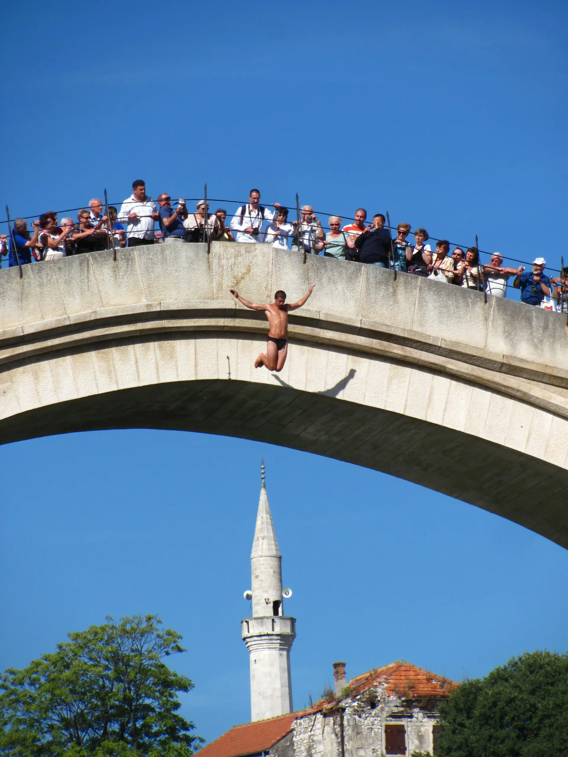 A diver in mid-air below the arch of the Stari Most, the crowd watching from the parapet