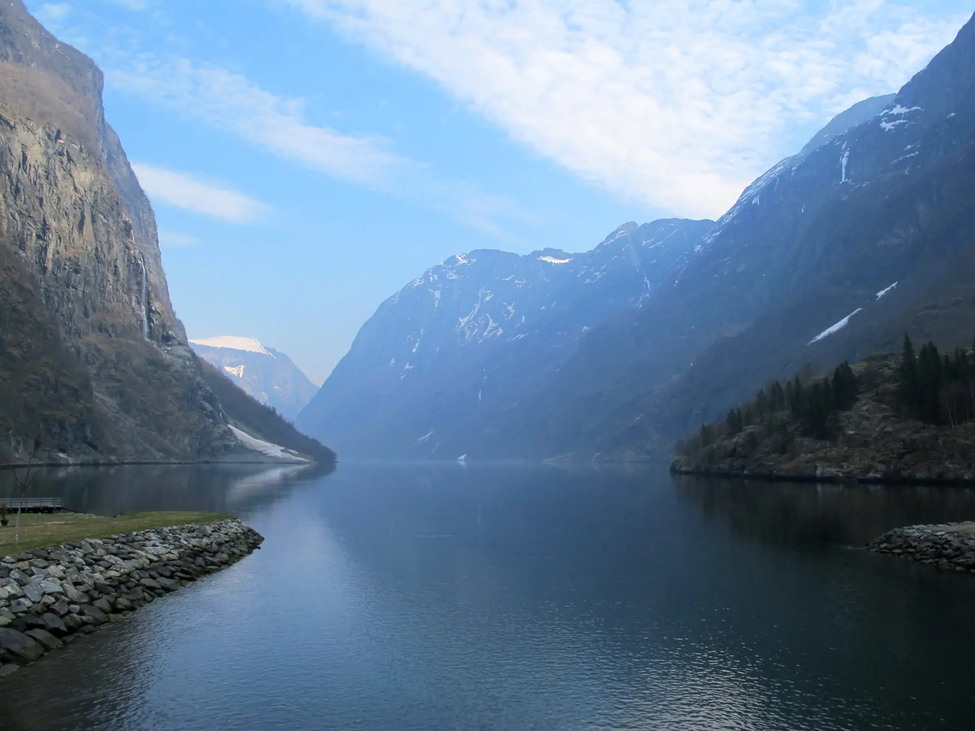 The narrow Nærøyfjord between steep snow-streaked mountain walls, calm water and a small boat far below