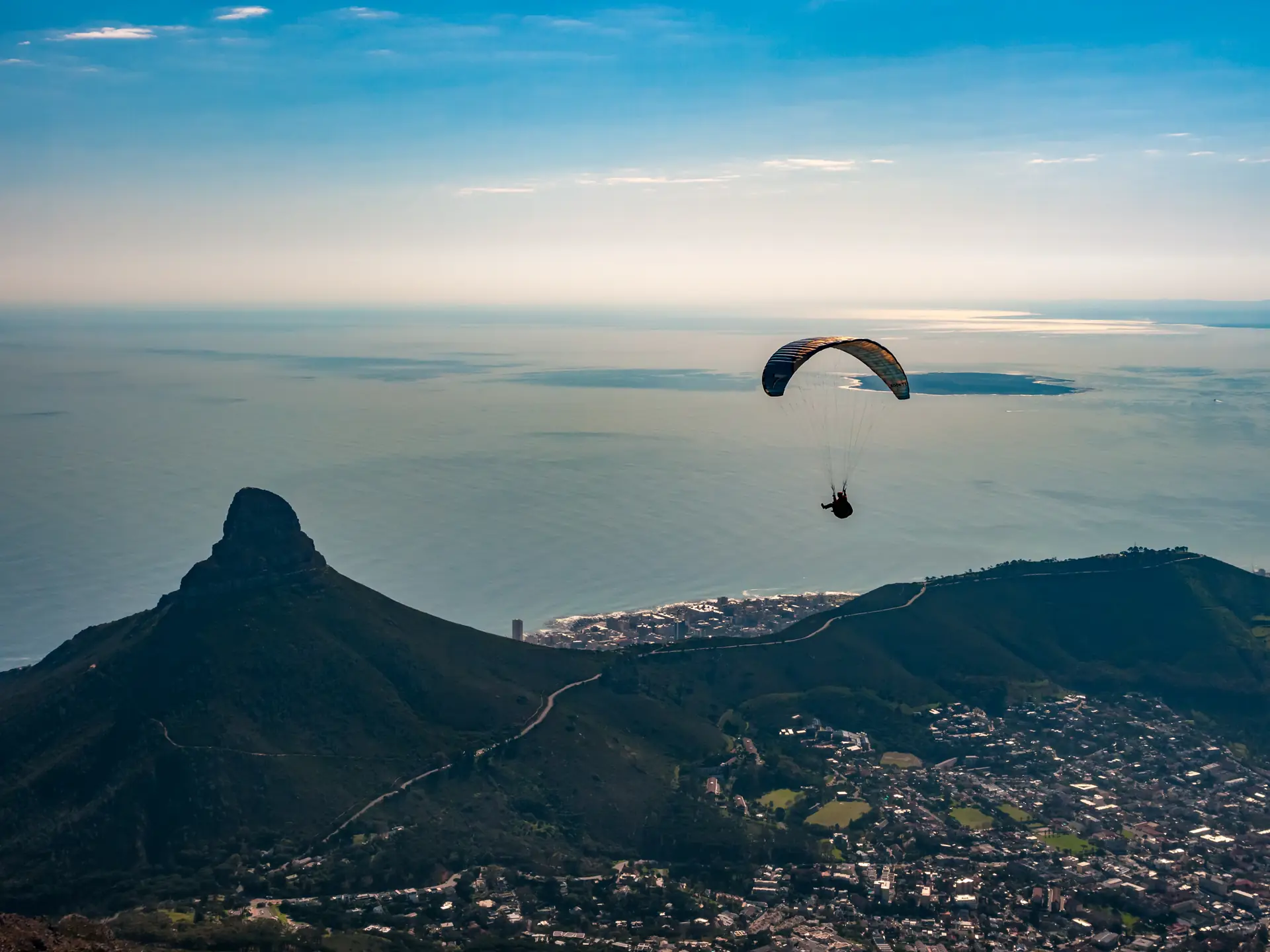 A tandem paraglider in the air above the slopes of Signal Hill with Lion's Head and the Atlantic coast beyond