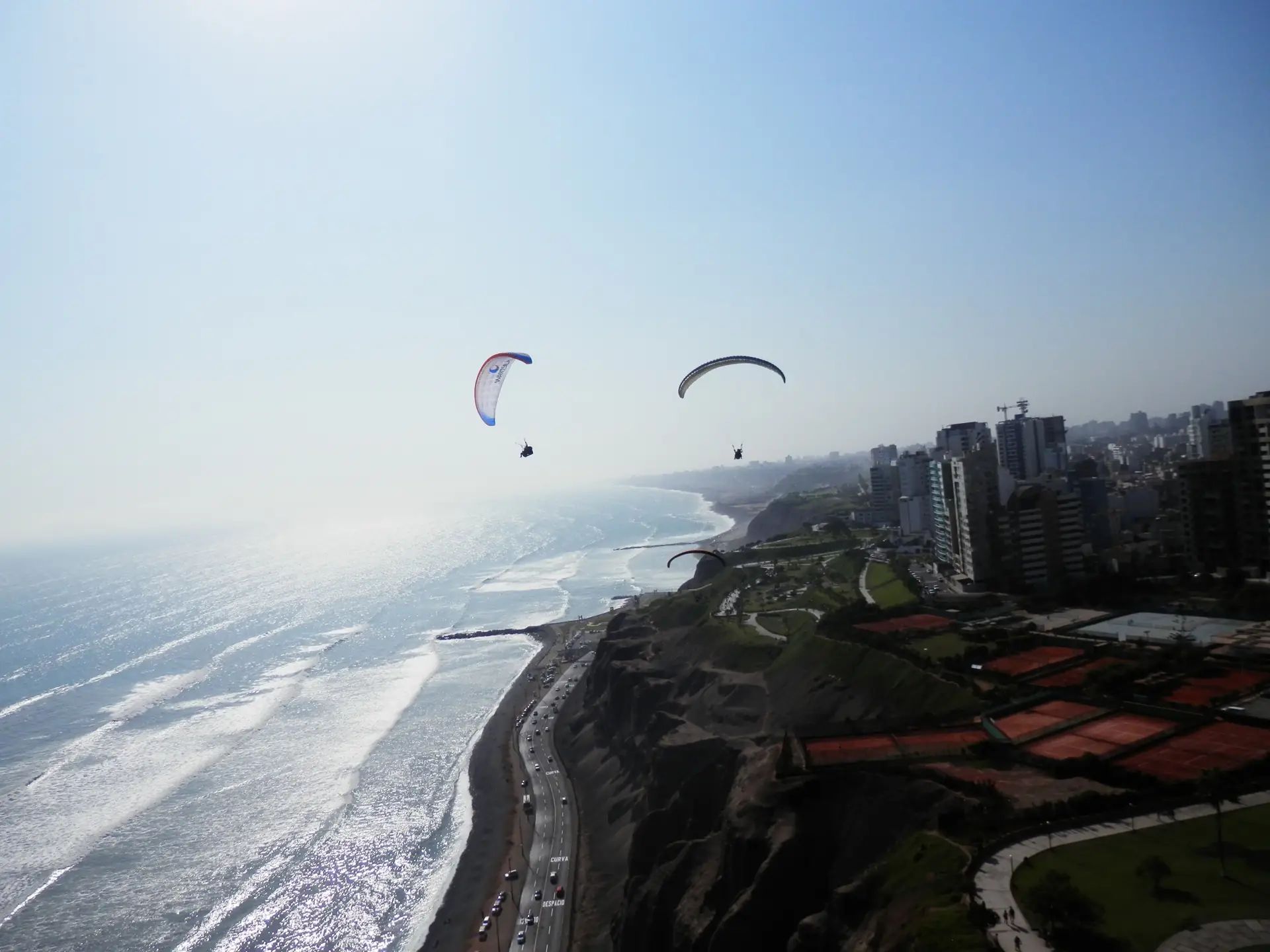 Zwei Gleitschirme über den Klippen von Miraflores in Lima, darunter die Küstenstraße, die Brandung und die Hochhäuser der Stadt