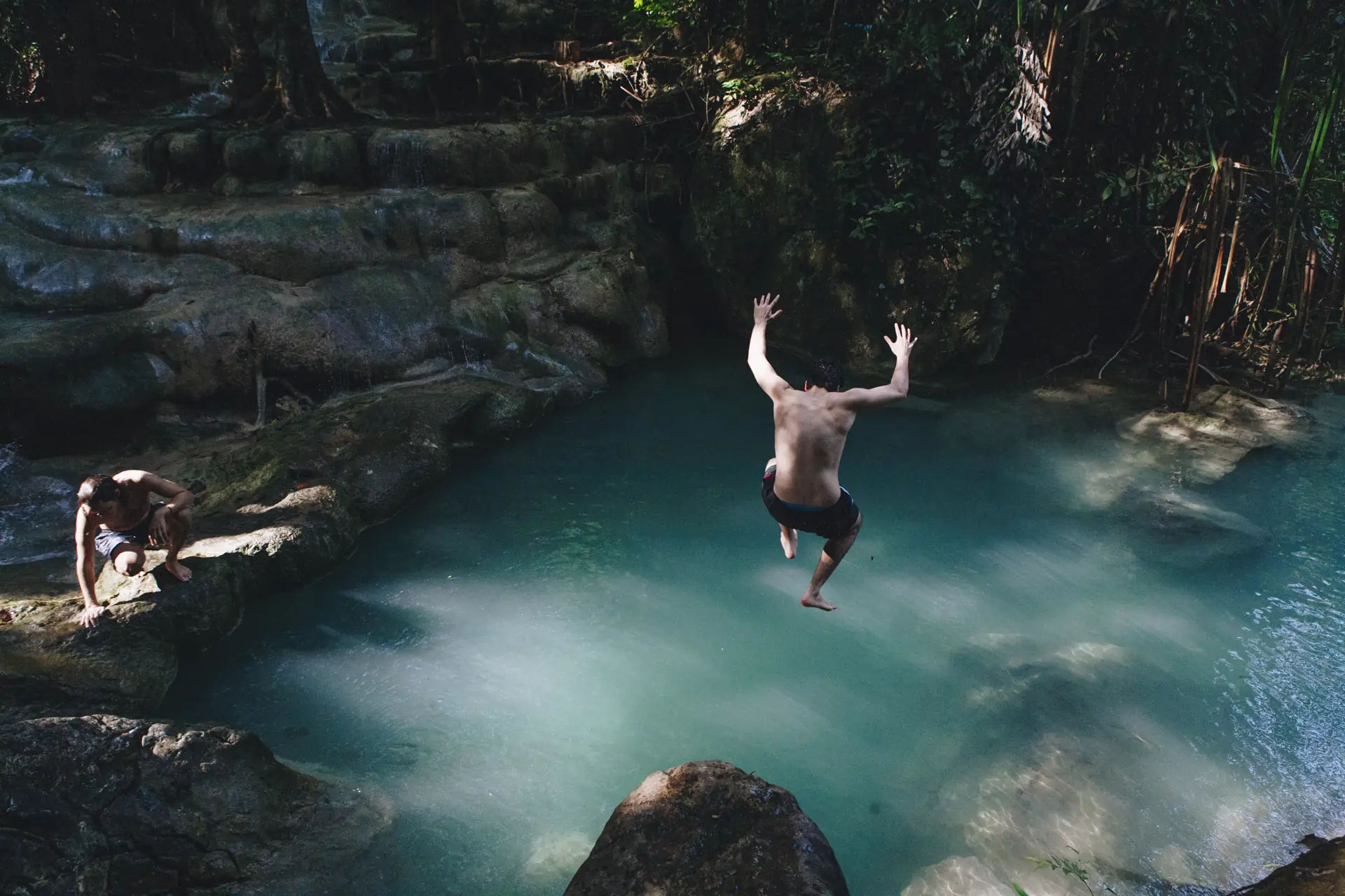 A man mid-air, arms up, jumping from a rock ledge into a turquoise jungle pool (symbolic image, not Kawasan)