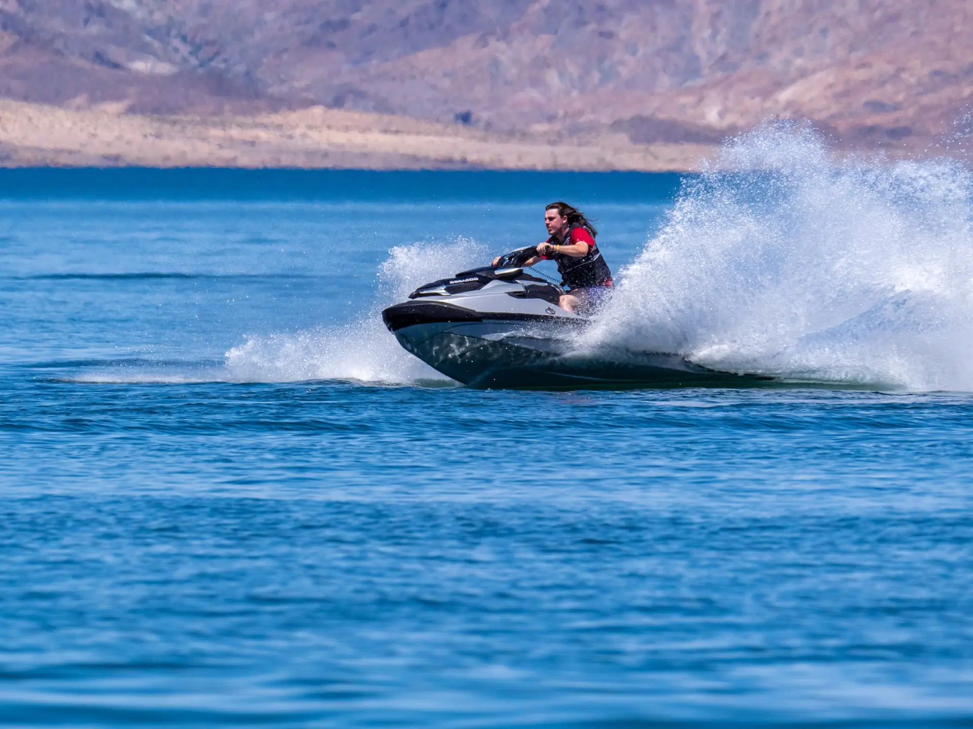 Ein Jetski-Fahrer in Schwimmweste zieht eine enge Kurve auf dem blauen Wasser des Lake Mead bei Boulder Harbor, Gischt fliegt, Wüstenhügel dahinter