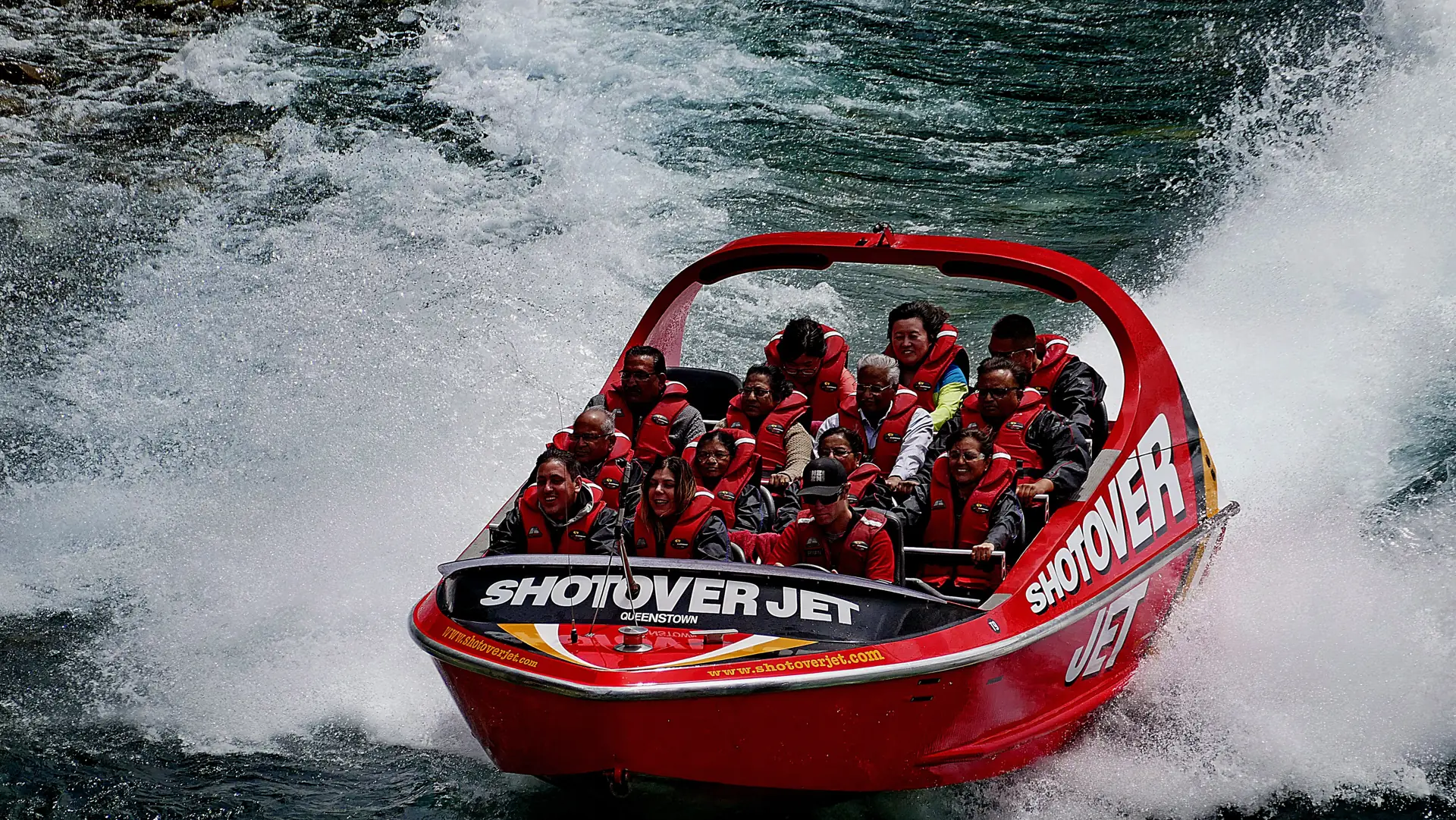 A red Shotover Jet boat full of passengers throwing up spray as it powers through the river canyon