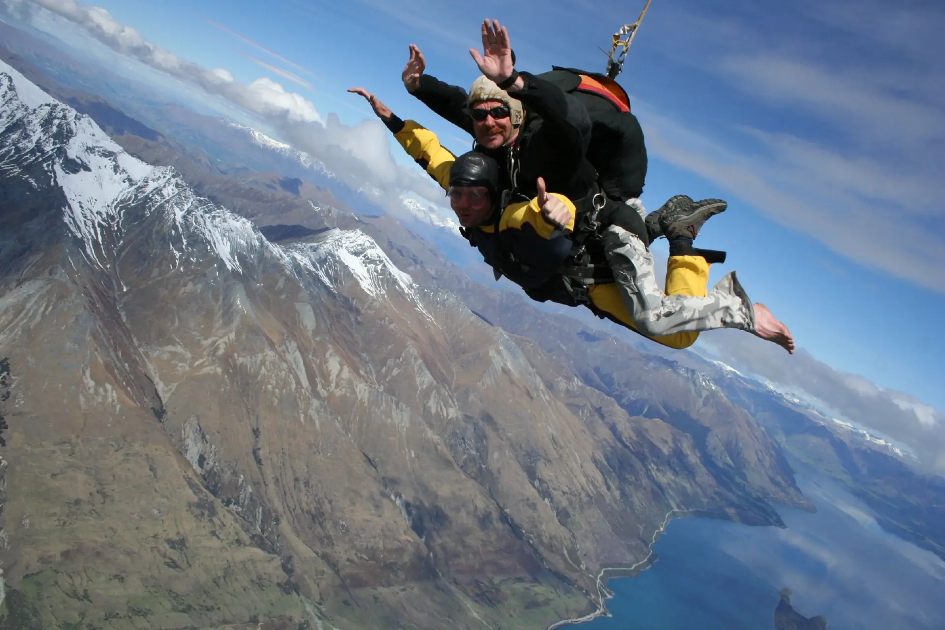 A tandem skydiving pair in freefall over the snow-streaked mountains and lake near Queenstown