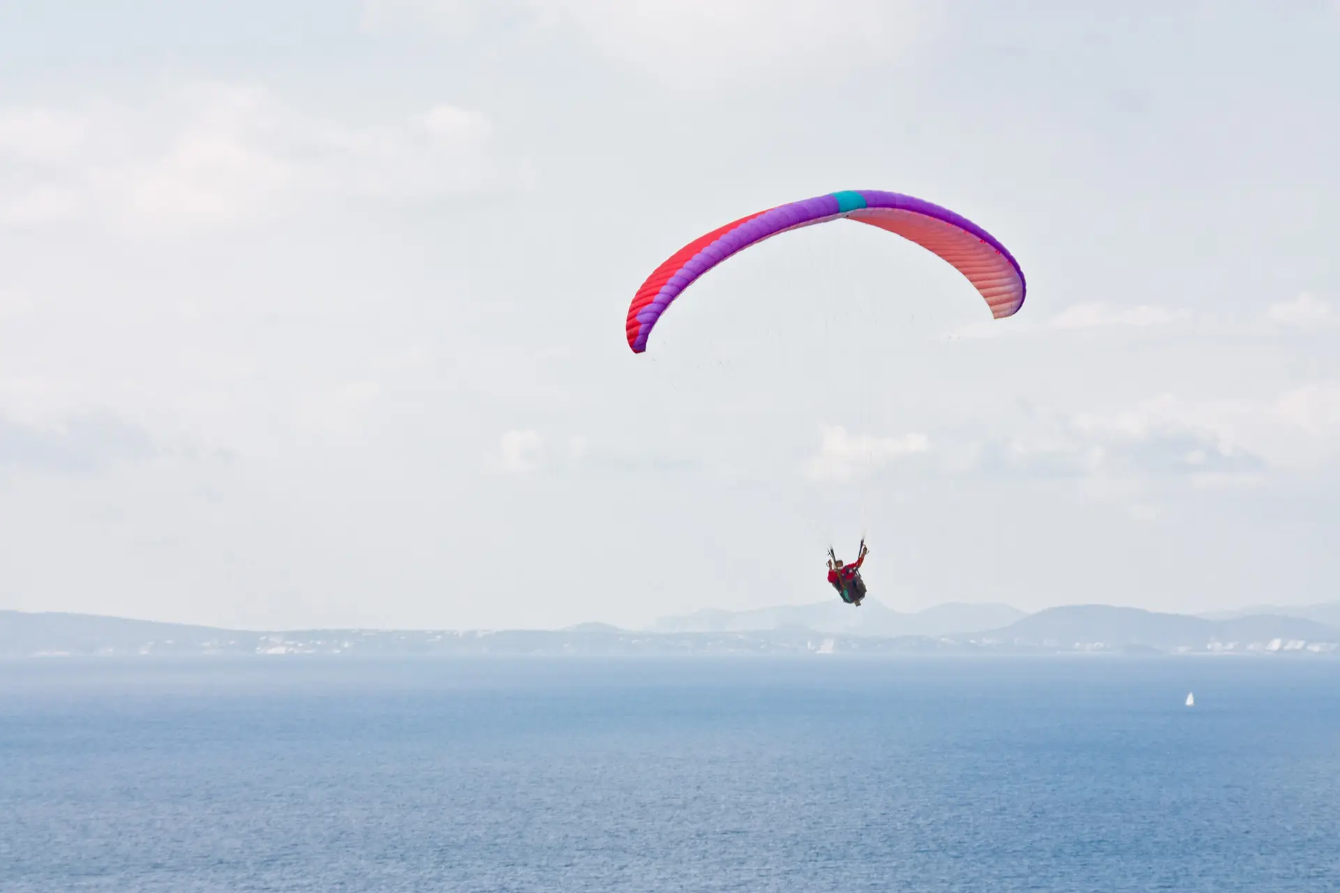 A tandem paraglider with a purple wing high over the sea off the Llucmajor coast of Mallorca, the horizon and a sailing boat below
