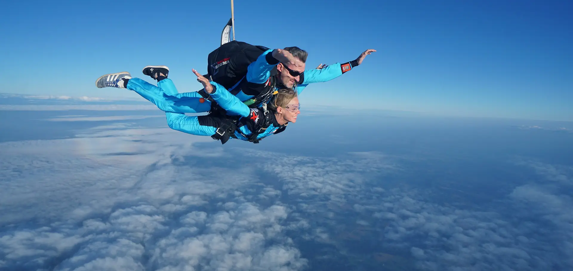 A tandem skydiving pair in freefall above a layer of cloud, the instructor behind the passenger
