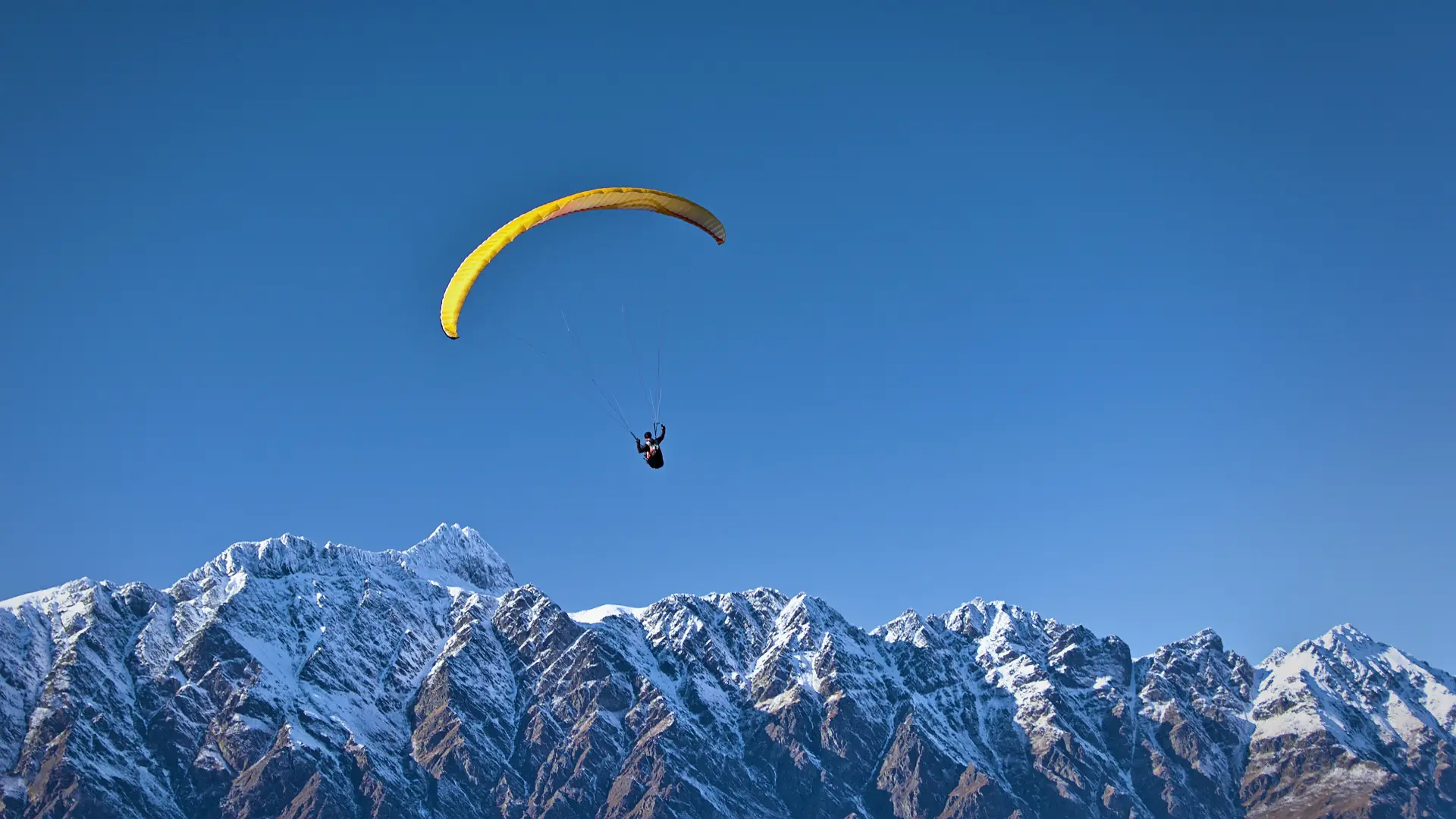 A yellow tandem paraglider soaring high above the snow-covered Remarkables range near Queenstown