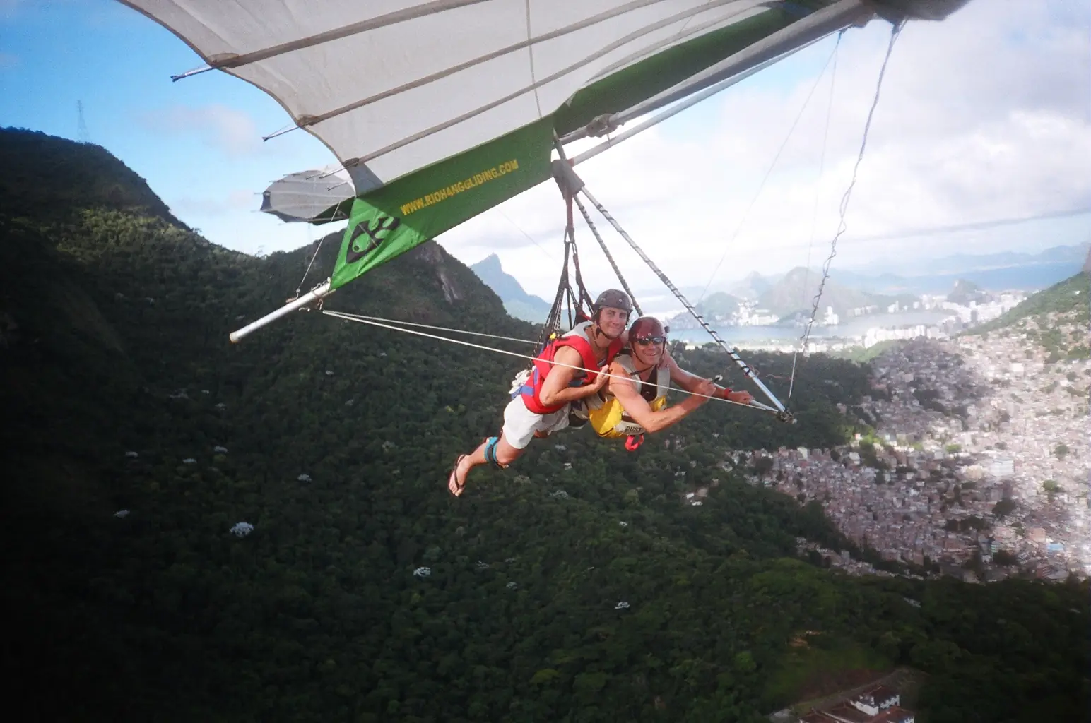 Ein Tandemdrachen im Flug, Pilot und Passagier liegen nebeneinander unter dem Flügel über dem Wald und der Stadt Rio de Janeiro (Symbolbild, Rio ist keines dieser Reviere)