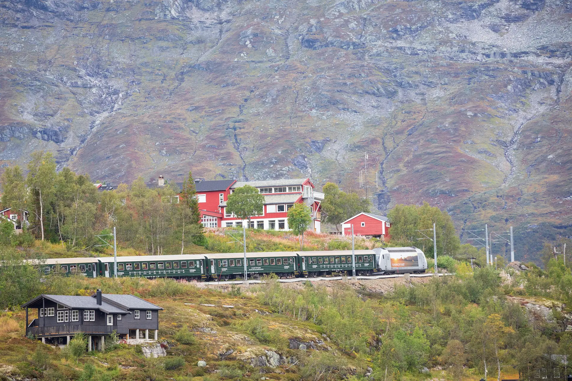 A green Flåm Railway train at the Vatnahalsen halt high in the mountains, the hotel and the Rallarvegen track beside it