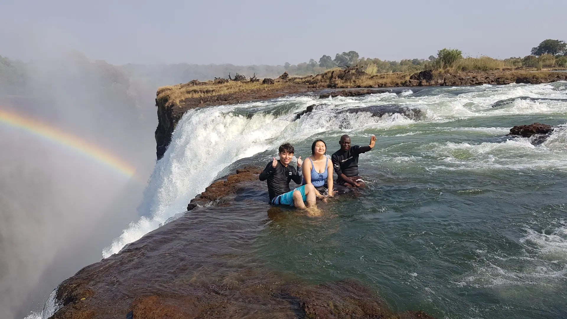 Swimmers sitting in Devil's Pool on the very lip of Victoria Falls with the water dropping away beside them