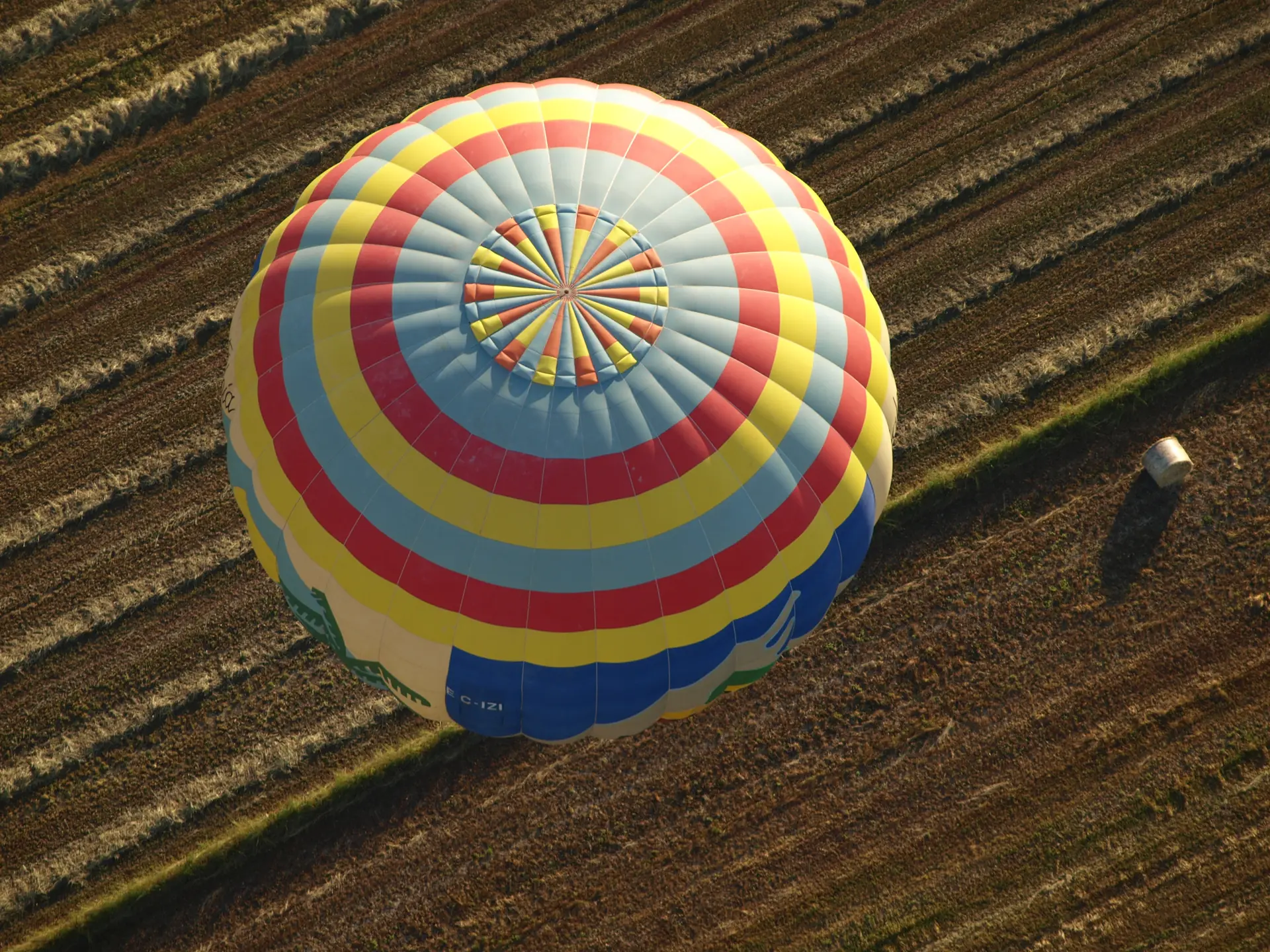 A striped hot-air balloon seen from above, drifting low over a ploughed field near Manacor with a person watching from the furrows